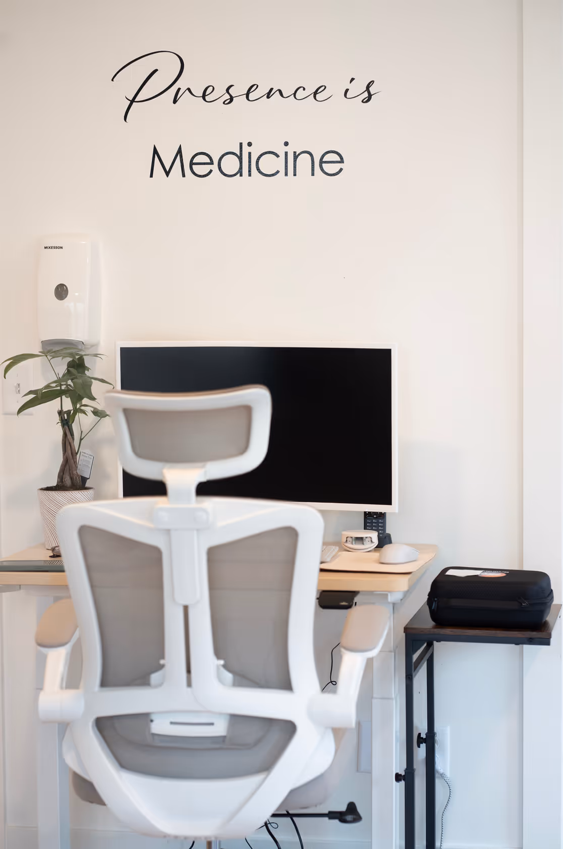 A white desk chair and monitor in a consultation room, below the words Presence is Medicine on the wall