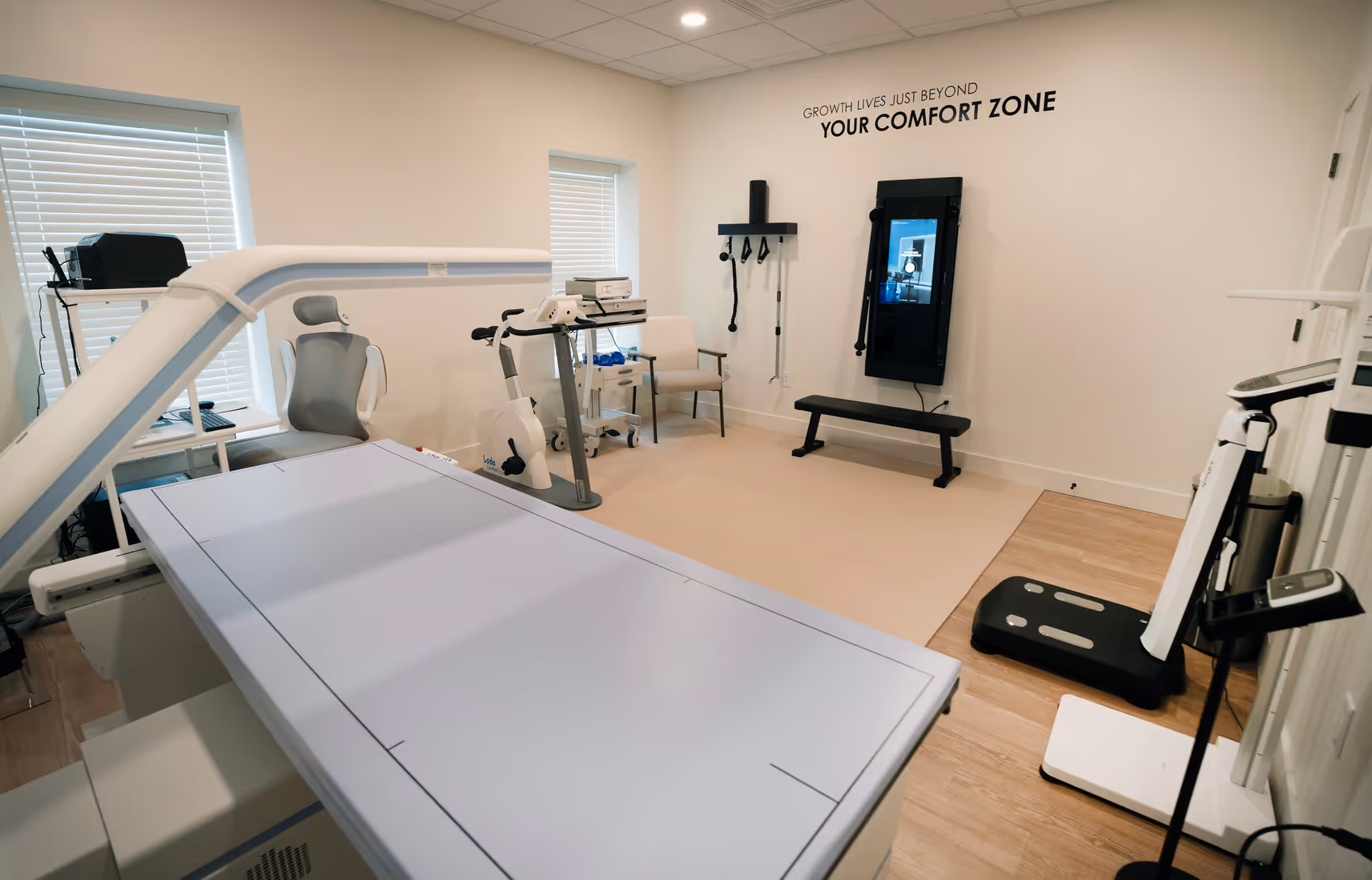 The testing room with a DEXA table, a stationary bike, and a wall-mounted fitness screen